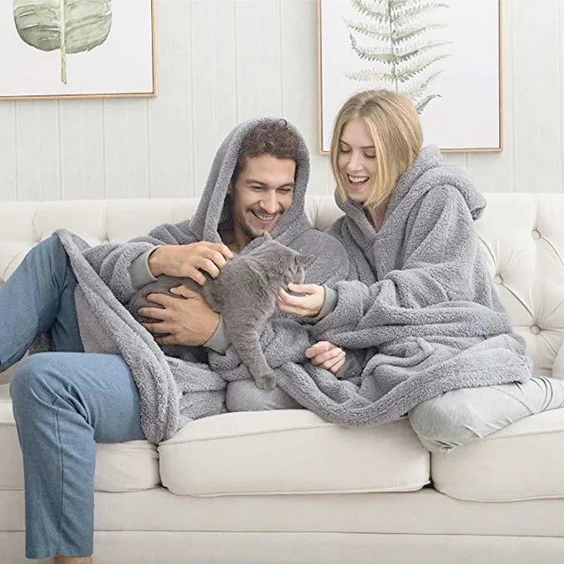 Two people sitting on a couch with a gray blanket wrapped around them, holding a cat.