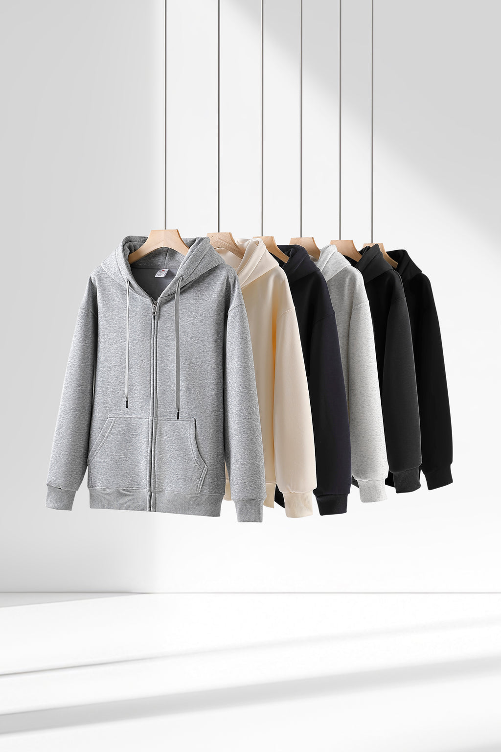 Six zip-up hoodies in different colors (gray, cream, black, white, dark gray, and black) hanging on wooden hangers against a clean white background with soft shadows.